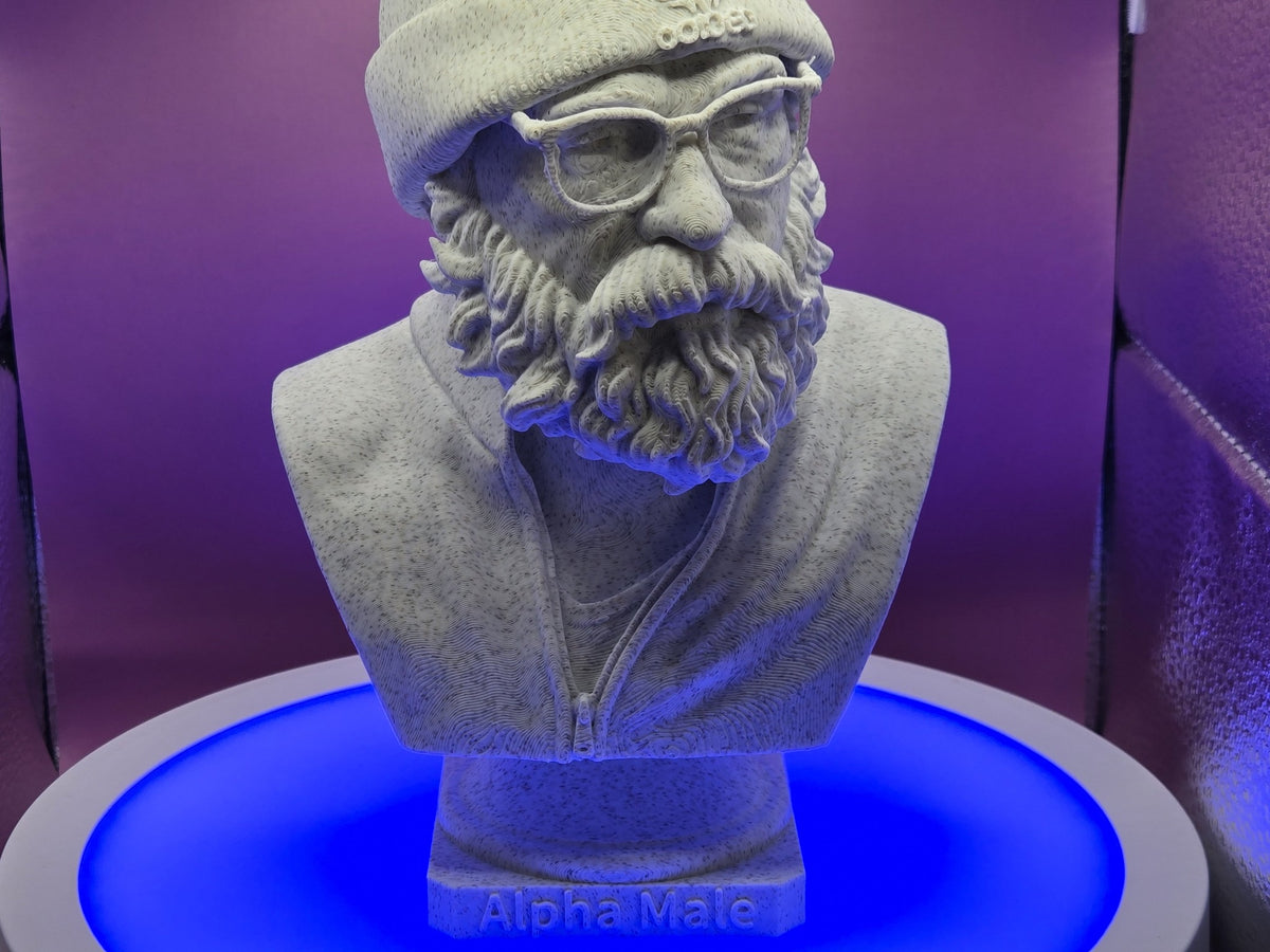 Alpha Male Bust by Mega Diamonds, real photo of the finished Marble PLA filament print on a blue-lit turntable, wide front view of the bearded bust in a beanie and glasses on a plinth lettered Alpha Male